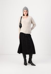 Maglione beige a maglia con scollo a V, gonna nera a trapezio con cuciture a contrasto, stivali neri a punta al ginocchio e berretto di maglia grigio.