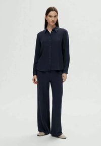 Mulher em pé, vestindo uma camisa de botão de manga longa azul marinho e calças largas combinando, com sapatos planos bege.