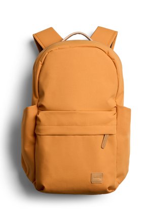 Sac à dos jaune moutarde avec bretelles rembourrées, poche frontale zippée, poches latérales et une petite étiquette de marque en bas à droite.