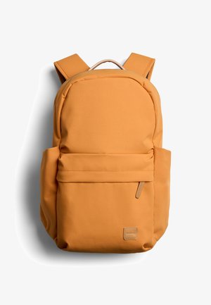 Sac à dos jaune moutarde avec bretelles rembourrées, poche frontale zippée, poches latérales et une petite étiquette de marque en bas à droite.