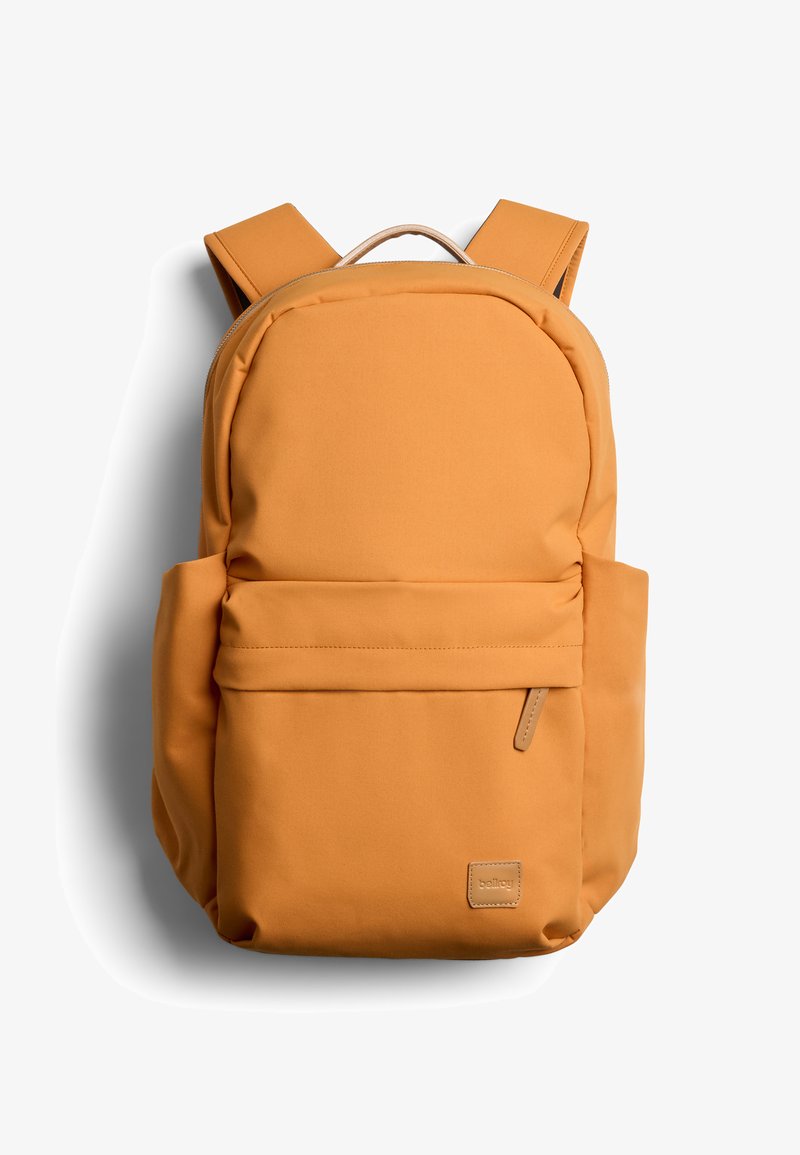 Zaino giallo senape con spallacci imbottiti, tasca frontale con zip, tasche laterali e una piccola targhetta del marchio nell'angolo inferiore destro.