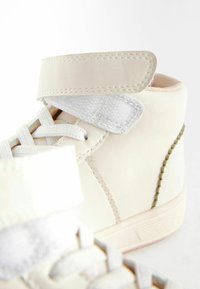 Baskets montantes blanches avec une surface en cuir lisse, des accents cousus, une sangle en Velcro en tissu et des lacets. Doublure intérieure rose pâle. Forme de bout arrondi.