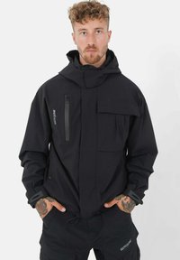 Giacca nera con cappuccio, dotata di numerose tasche, polsini regolabili e tasca sul petto con zip. Realizzata in tessuto liscio e resistente.