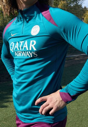 Personne portant un maillot d'entraînement de football Paris Saint-Germain bleu canard et violet avec le logo Qatar Airways, debout sur un terrain herbeux près d'un filet de but.