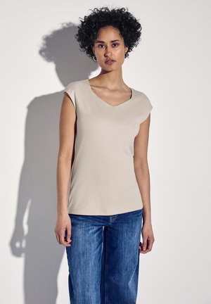 T-shirt beige a V con maniche corte, tessuto morbido, forma leggermente aderente, abbinato a jeans di denim larghi. Ombra lieve sulla parete.