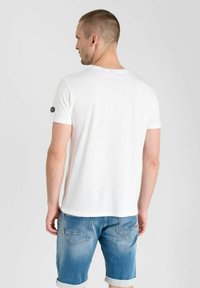 Camiseta blanca de manga corta con escote redondo y un acento de logo negro en la manga izquierda, combinada con pantalones cortos de mezclilla azul. Textura de tela suave.