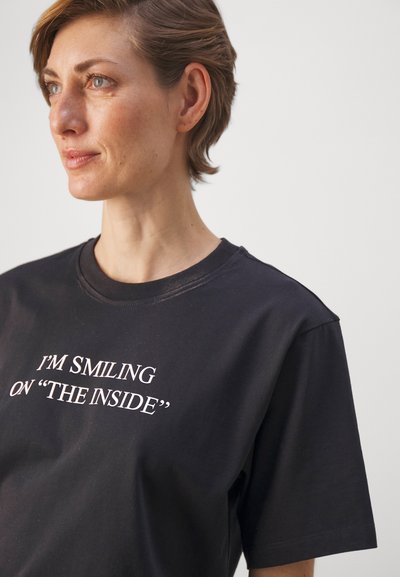 Musthave musta T-särk lühikeste varrukatega ja ümaratest, millel on valge tekst "NAERATAN SISELT".