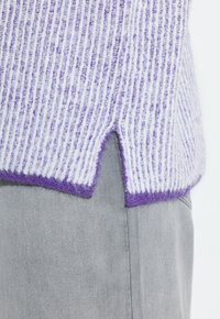Maglione lavorato a maglia con strisce verticali viola e bianche, caratterizzato da un orlo spaccato e rifiniture viola. Indossato sopra un tessuto grigio.