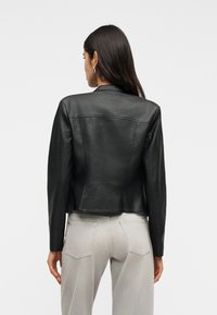 Γυναίκα με μακριά σκούρα μαλλιά ντυμένη με εφαρμοστή μαύρη δερμάτινη ζακέτα και ανοιχτό γκρι τζιν, φαίνεται από πίσω με φόντο απλό.