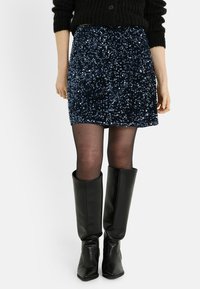 Mini falda azul marino con lentejuelas, de diseño ajustado, con una textura suave y superficie brillante, combinada con botas negras hasta la rodilla.