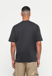 Mand med kort hår iført en ensfarvet mørkegrå T-shirt og beige cargo-bukser, vendt væk mod en ensfarvet hvid baggrund.