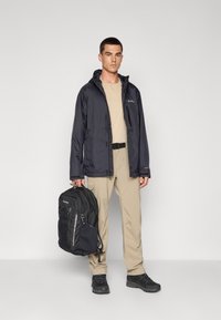 Veste imperméable noire, t-shirt beige, pantalon cargo beige, sac à dos noir, chaussures noires, se tenant devant un fond uni.