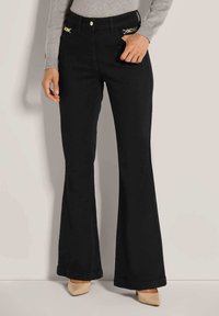 Frau in schwarzen Schlaghosen mit goldenen Kettendetails an den vorderen Taschen und beigen spitzen Absatzschuhen, die vor einem schlichten Hintergrund steht.