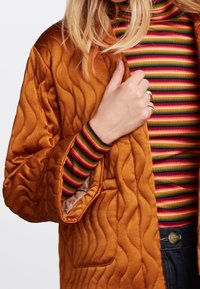 Kobieta ubrana w brązową pikowaną kurtkę na kolorowy, paskowany golf, trzyma kurtkę w okolicy kołnierza, mając na palcach pierścionki.