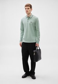 Polo de manga comprida verde-claro com detalhes em preto na gola, combinado com calças pretas e bolsa de couro preta. Textura suave e corte ajustado.