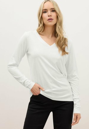 Femme blonde aux cheveux ondulés portant un haut blanc à manches longues avec un col en V et un pantalon noir, posant avec une main dans la poche.