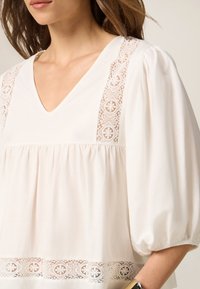 Blusa color crema con escote en V, mangas abullonadas y detalles de encaje a lo largo de los hombros y el dobladillo. Tejido suave con un acabado texturizado.