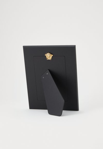 Czarna prostokątna ramka na zdjęcia z pochylonym stojakiem i małym złotym emblematem dekoracyjnym na środku u góry na białym tle.