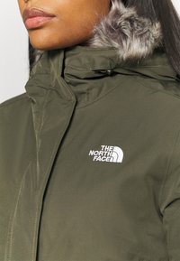 Oliwkowa kurtka z futrzanym kapturem. Charakteryzuje się przeszytymi szwami oraz białym haftowanym logo na klatce piersiowej. Gładki, trwały materiał.