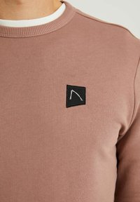 Sudadera de algodón en color malva con un cuello de canalé, que presenta un parche rectangular negro con un diseño geométrico blanco. Textura suave, diseño casual.