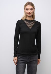 Schwarzes Langarmoberteil mit einem V-förmigen Spitzenpaneel am Ausschnitt, mit gewelltem Rand und figurbetontem Design. Kombiniert mit dunklen Jeans.