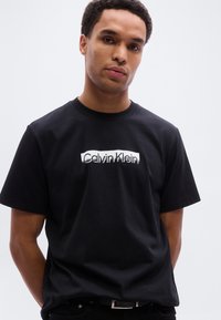 Μαύρη βαμβακερή μπλούζα με λευκό λογότυπο "Calvin Klein" στο κέντρο. Στρογγυλή λαιμόκοψη, κοντά μανίκια και κλασική εφαρμογή.