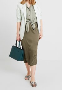 Nainen, joka käyttää vaaleaa bleiseriä, sidottua oliivinvihreää paitaa, yhteensopivaa hameetta, kantaa teal-väristä käsilaukkua ja käyttää kuvioituja liukuvaatteita, seisoo yksinkertaisella taustalla.