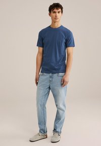 Majica s kratkimi rokavi v modri barvi iz mehkega bombaža, v kombinaciji z svetlo modrimi kavbojkami in sivi obutvi, prikazana na modelu na nevtralni podlagi.