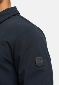 Dunkelblaue Jacke mit glatter Textur, strukturierter Kragen und schwarzem Gummilogoklett auf der Schulter.