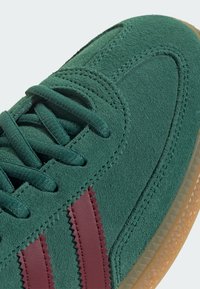 Zapatilla de ante verde con rayas marrón, parte superior texturizada y suela de goma. Presenta cordones planos y costuras en zigzag.