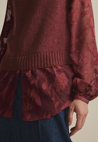Pull en tricot bordeaux avec un ourlet côtelé superposé sur un blouse transparente brodée présentant des motifs floraux et une fermeture à boutons.