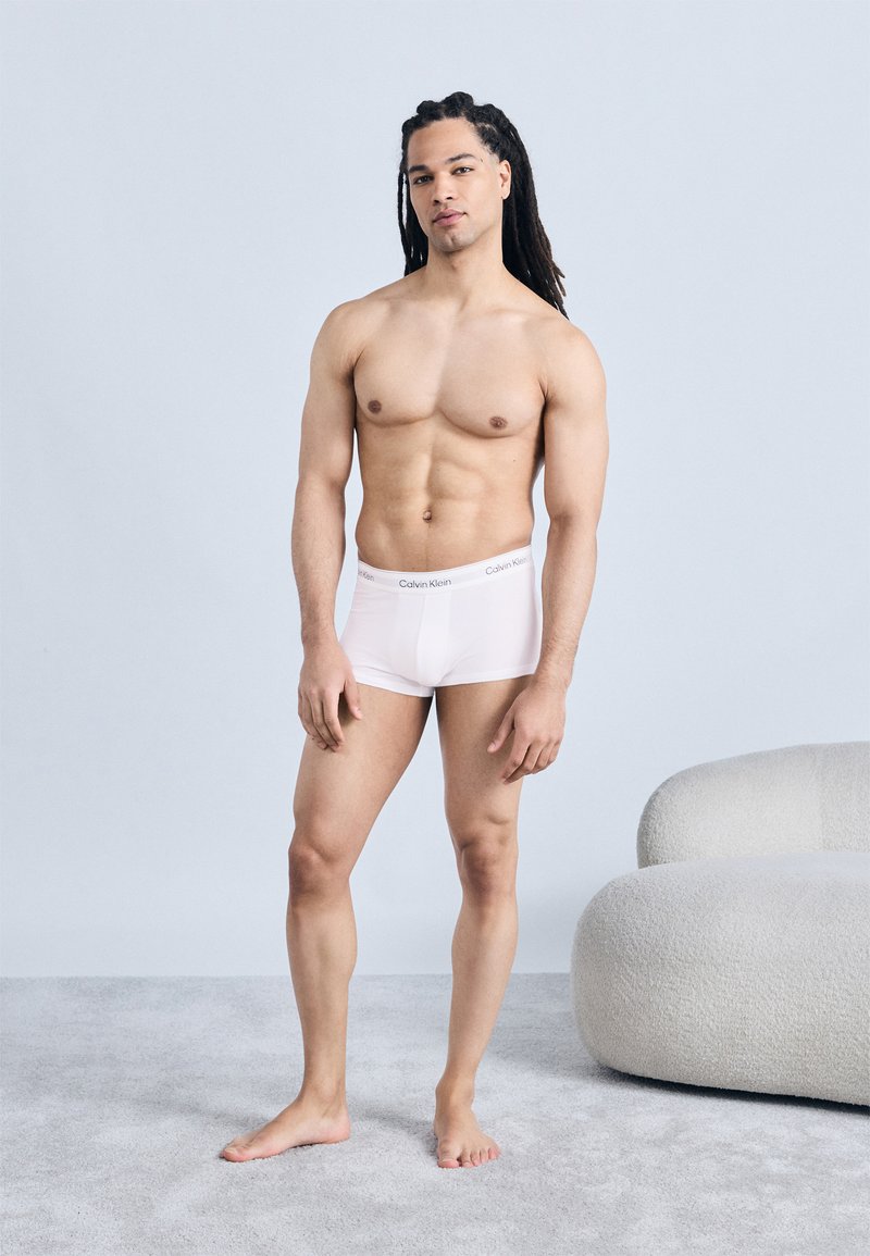 Νεαρός άνδρας με μακριά πλεξούδα στέκεται ξυπόλητος σε γκρι χαλί φορώντας λευκά εσώρουχα Calvin Klein κοντά στους στρογγυλεμένους λευκούς πολυθρονιού.