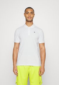 Hvit polo skjorte med knappelukking i to knapper og korte ermer, med en liten svart logo på brystet. Båret med lysegule shorts.