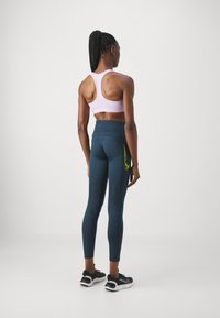 Soutien-gorge de sport rose clair et legging vert foncé avec des accents jaunes, tissu texturé, design ajusté, et chaussures de sport avec une tige noire.