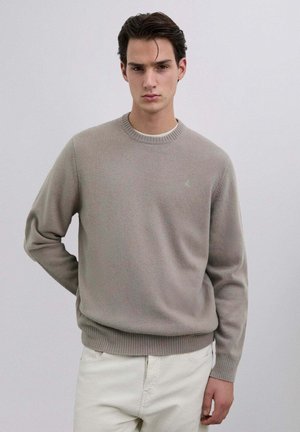 Suéter de punto gris claro con cuello redondo, puños y dobladillo acanalados, con un logo sutil en el pecho. Combinado con pantalones de color claro.