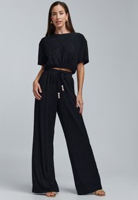 Černý dvoudílný outfit se zkráceným, texturovaným topem a volnými kalhotami. Mezi výrazné detaily patří stahovací šňůrka se ozdobnými korálky.