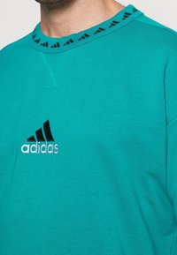 Sweat-shirt bleu sarcelle avec un col rond orné d'un logo noir et d'une bordure à motifs au niveau du col. Texture douce en coton, coupe décontractée.