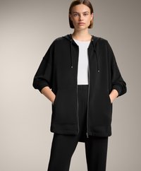 Felpa nera con zip realizzata in tessuto spesso, dal taglio ampio, con tasca frontale e cordini regolabili, abbinata a pantaloni neri.