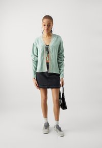 Mintgrønn cardigan med knytefront og broderte detaljer, kombinert med et svart denimskjørt og grå sneakers. Svart håndveske.