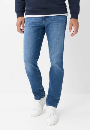 Mand iført blå denimjeans, hvide sneakers og en mørk langærmet sweatshirt står mod en ensfarvet baggrund.
