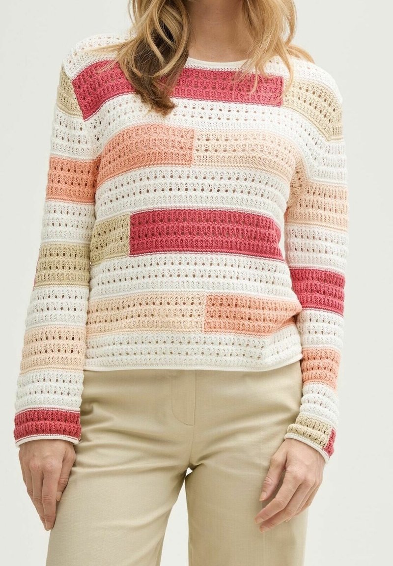 Femme portant un pull en crochet multicolore à manches longues avec un pantalon beige, debout devant un fond clair uni.