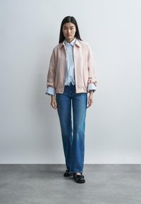 Ung kvinde står iført en pink jakke over en stribet skjorte, blå jeans og sorte loafers mod en ensartet lys baggrund.