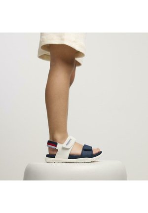Sandalias azules y blancas con correas ajustables, suela de goma y un diseño texturizado. Exhibidas sobre un pie en pedestal para mayor visibilidad.