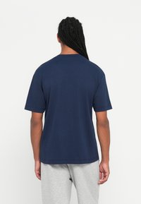 Άτομο με μακριά πλεγμένα μαλλιά στέκεται με την πλάτη, φορώντας ναυτική μπλε κοντομάνικη μπλούζα και ανοιχτό γκρι παντελόνι σε απλό φόντο.