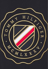 Logo Tommy Hilfiger, který zobrazuje štít s červenými, bílými a černými pruhy, rámovaný zlatou barvou, na tmavém pozadí. Okolo štítu je vyšitý text.
