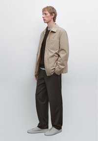 Beige jakke med trykknapper, båret over en brun struktureret sweater, parret med mørke bukser og lysegrå sneakers på en neutral baggrund.