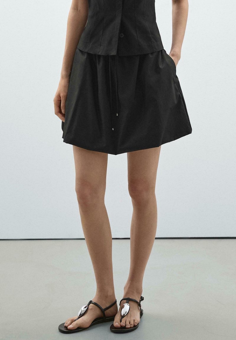 Gonna nera di cotone con vita regolabile tramite coulisse, abbinata a sandali neri bassi con dettaglio in metallo. Design semplice e moderno con vestibilità comoda.