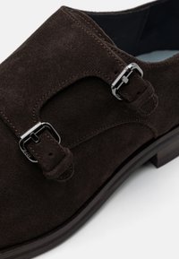 Chaussure en daim marron avec un détail de boucle et des coutures contrastées. Le design possède un bout rond et un talon en bois.