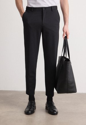 Pantaloni neri con texture, dal taglio slim, dotati di bottone frontale e tasche laterali, indossati con scarpe in pelle nere e una borsa a tracolla nera.