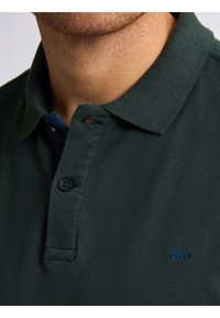 Donkergroen poloshirt van gestructureerde stof. Heeft een kraag, twee knopen en een klein blauw logo op de borst. Close-up weergave.